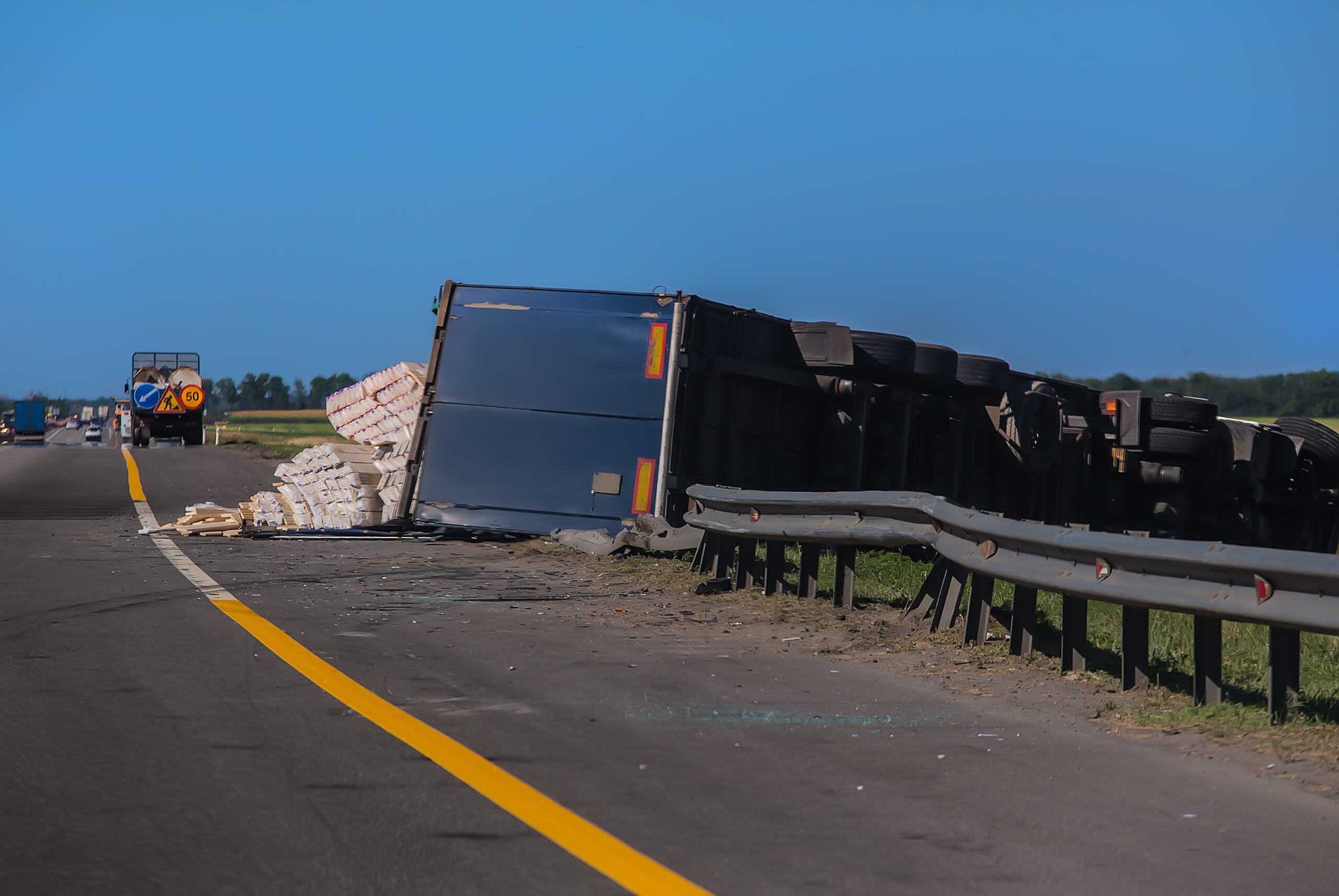 A large commercial semi-truck trailer flipped completely onto its side on the edge of a highway, crushing a metal guardrail and spilling its cargo, showcasing a severe crash managed by Pensacola jackknife truck accident lawyers.